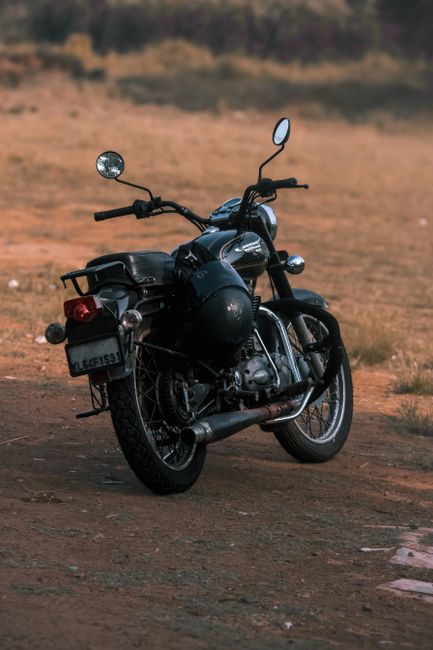 A black cruiser-style motorcycle with chrome accents and a leather seat is parked on a dirt surface in an open, rural area with dry grass and sparse vegetation in the background. The motorcycle is equipped with rearview mirrors, a small windscreen, and a rear luggage rack. A black helmet is securely placed on the seat, and protective plastic wrap is visible around some parts of the motorcycle, indicating it may be in transit or awaiting pickup. The lighting suggests early morning or late afternoon, with soft natural light illuminating the scene. The setting lacks other vehicles or people, emphasizing the motorcycle's stationary position. This image is relevant to home relocation and packing and moving services, reflecting the transportation aspect of furniture and belongings during a property move, as handled by companies like Man with Van Park Royal, especially when considering local regulations such as Brent Council's bulky waste rules affecting move logistics in the Park Royal area.