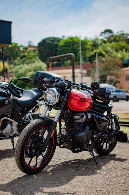 A black and red motorcycle, parked on a concrete surface outdoors, with a large round headlamp, black handlebars, and a red fuel tank featuring the Royal Enfield logo. The motorcycle is positioned alongside another motorcycle, which is partially visible to the left. In the background, there are trees, a building, and a parking lot with several cars and vans under a partly cloudy sky. The scene is well-lit, indicating daytime, and the motorcycle appears ready for transport or part of a home or vehicle relocation process. This image aligns with house removals and moving logistics, illustrating the careful handling and transportation of personal vehicles during home moves, as offered by Man with Van Park Royal.