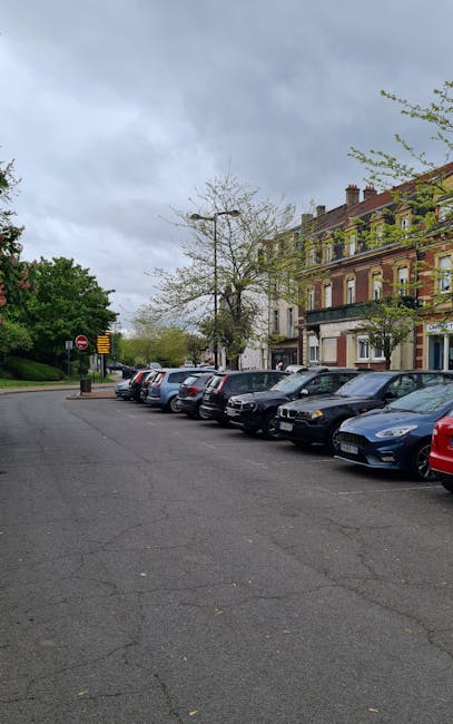 A row of parked cars along a residential street with pavement, some new or recently cleaned, under an overcast sky. The vehicles include various sizes and colours, parked adjacent to a multi-storey brick building with commercial establishments on the ground floor. In the background, there are leafless and lush green trees, indicating early spring or late autumn. The street is compliant with parking regulations, with a clear no-entry sign visible at the end. This parking arrangement is suitable for house removals and furniture transport, supporting logistic planning for a home relocation. Occasionally, Man with Van Park Royal might use this area for loading or unloading household furniture and boxes during local removals, although no activity is visible in this particular image.