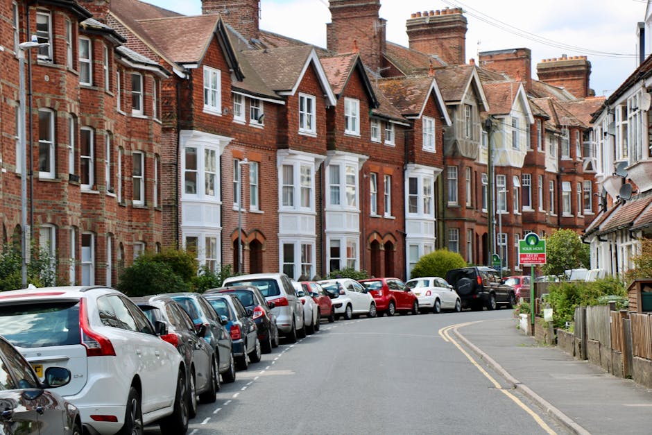 A residential street scene featuring a row of traditional brick houses with pitched roofs and white window frames, situated along a curved, narrow road. Numerous parked cars of various makes and models are lined up along both sides of the street. In the foreground, a white van is partially visible, likely involved in a home relocation or furniture transport process, with a man with van service such as Man with Van Park Royal performing a loading or unloading task. The pavement is clear of obstructions, and there is a parking permit sign visible on a lamppost. The environment is well-lit with natural daylight, highlighting the details of the houses, cars, and surrounding greenery, reflecting typical surroundings where house removals and packing logistics occur in the context of local moving services. The scene captures the urban atmosphere of Park Royal, London, ideal for illustrating house removals and moving logistics.