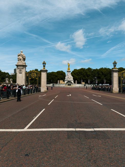 A clear view of a large, open paved area leading up to an ornate entrance gate with four tall stone pillars, each topped with decorative statues and lanterns. Behind the gate, a statue with a golden figure stands atop a white pedestal, surrounded by a line of trees and lush greenery. To the left and right sides of the paved area, groups of people are gathered, some standing and others seated, creating a scene of social activity. The paved space features white painted lines indicating parking spaces, with one small vehicle visible in the distance near the gate. The sky above is mostly clear with a few wispy clouds and bright sunlight illuminating the scene. This setting, captured outdoors, reflects an urban environment possibly near a historic monument or park, illustrating the importance of orderly traffic management and pedestrian space.