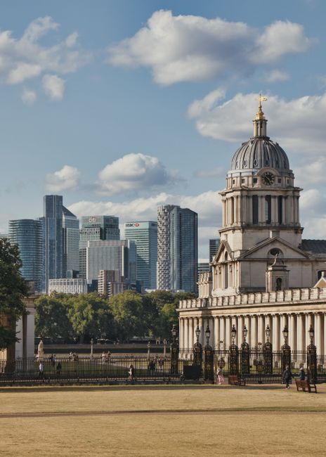 The image shows a historic, ornate domed building with classical architectural features, including tall columns and decorative stonework, situated in a park with a well-maintained lawn and a black wrought iron fence. In the background, there is a modern city skyline with high-rise office buildings made of glass and steel, under a partly cloudy sky with scattered clouds. The scene is illuminated by daylight, highlighting the contrast between the traditional structure and the contemporary cityscape beyond. This setting is indicative of London, with the historic building possibly being part of a government or cultural site. No moving or packing activities are visible in the image, consistent with a static urban environment.