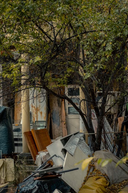 An outdoor scene showing a yard or alleyway filled with discarded household items and furniture, including a partially visible white kitchen cupboard, wooden cabinets, and various boxes wrapped in pla