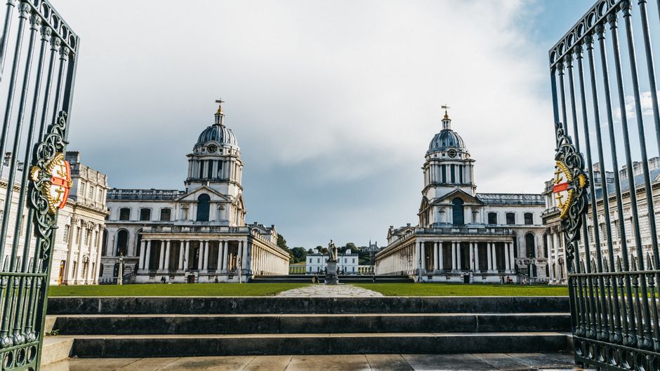 A symmetrical view of a historic neoclassical building complex, seen through an ornate black wrought iron gate with gold accents at the hinges, leading to wide stone steps ascending to the main entran
