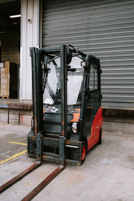 A compact electric forklift with a black protective cabin and red base is positioned outdoors on a concrete surface in front of a building with a partially open metal shutter door. The forklift's forks are lowered and extended forward, resting on the ground, ready for lifting or load transfer. Nearby, yellow parking lines are faintly visible on the pavement, indicating designated loading areas. The building features a brick ledge beneath the shutter, and an open interior space can be partially seen through the door opening. This forklift is equipped with hydraulic mechanisms used for lifting and transporting furniture, boxes, or appliances during home or commercial relocation processes. The scene captures the loading or unloading stage of a house removal, involving precise handling and transportation logistics, with the vehicle likely being used by a professional removals company such as Man with Van Park Royal, who specialise in home removals and furniture transport. The environment is well-lit with natural daylight, emphasizing the operational context for professional packing and moving services.