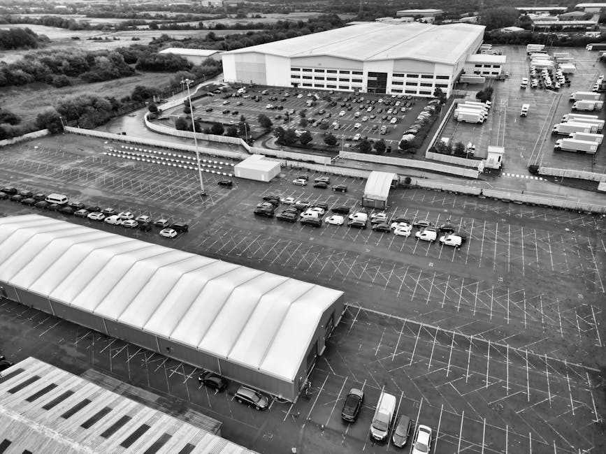 Aerial view of a large industrial warehouse situated within a commercial estate, with a surrounding extensive parking lot filled with numerous parked cars and vans. The warehouse has a white, pitched roof and multiple loading bays along its side, with some vehicles parked close to these bays, indicating active loading or unloading. Outside the warehouse, there are designated areas with clearly marked parking spaces and traffic routes, along with a curved access road leading to the estate. Adjacent to the facility, smaller structures, possibly storage units or site offices, are visible, along with a few trucks and delivery vehicles parked on the premises. The environment is well-lit with natural daylight, and the setting suggests a busy logistics hub engaged in home removals or furniture transport, visualized from high above to provide a comprehensive overview of the loading area, vehicle movements, and the scale of the site managed by companies like Man with Van Park Royal involved in packing and moving services, supporting efficient relocation logistics.