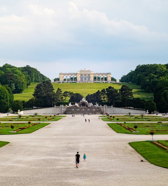 A wide outdoor scene showing a large, open gravel pathway leading towards a historic building situated atop a gently sloping grassy hill. In the foreground, a man and a young girl are walking along th