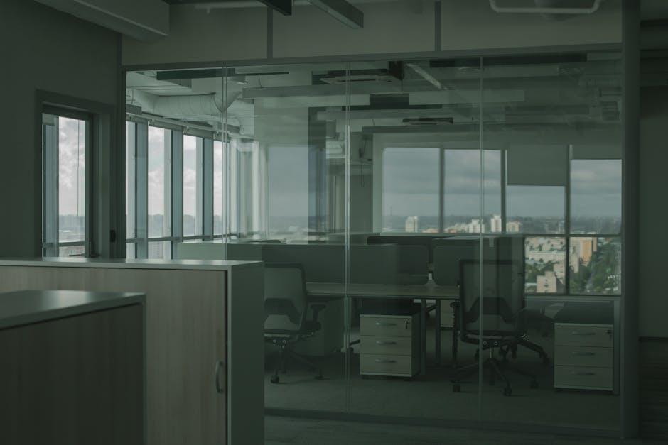 The image depicts an empty office space with glass walls and large windows that provide a view of a cityscape with buildings and cloudy sky outside. Inside, there are several workstations consisting of desks and office chairs with fabric upholstery. The desks are made of light-colored wood or laminate, and some are positioned against the glass walls, while others are facing inward. Small under-desk filing cabinets are visible next to some desks. The office has a modern, open-plan layout with white ceiling panels and exposed ceiling infrastructure, including ducts and support beams. Natural light illuminates the space, creating a bright environment suitable for a home relocation or office move. Although no moving equipment, boxes, or furniture in transit are visible in this particular shot, the setting suggests a professional backdrop for logistical activities like packing, furniture transport, or loading as part of a house or office removal service, such as provided by Man with Van Park Royal.
