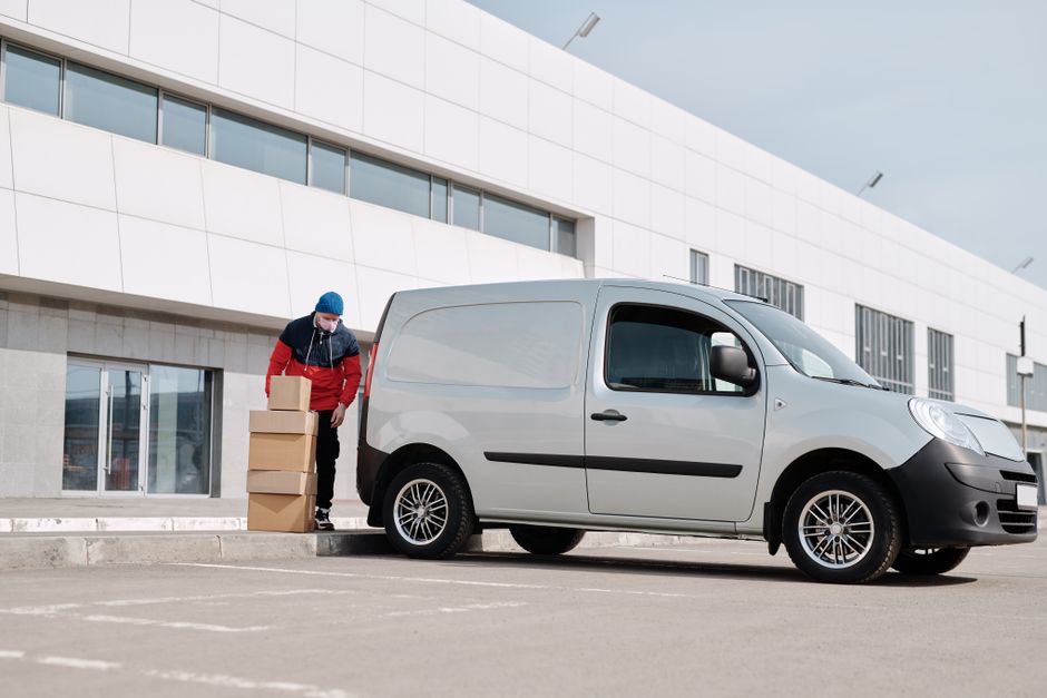 A man dressed in a red and black jacket, wearing a blue knit hat and a face mask, is standing outside a modern white building with large windows, lifting and stacking cardboard boxes near a small silver van parked on the paved pavement. The van is a compact commercial vehicle with black side mirrors, black wheel arches, and a sliding side door, positioned close to the building’s entrance. The scene captures a home relocation or furniture transport process, with the man preparing boxes for loading or unloading as part of a professional removals service provided by Man with Van Park Royal. The environment is well-lit with natural daylight, appearing to be a typical urban or business area facilitating packing and moving activities within the context of house removals or moving logistics.