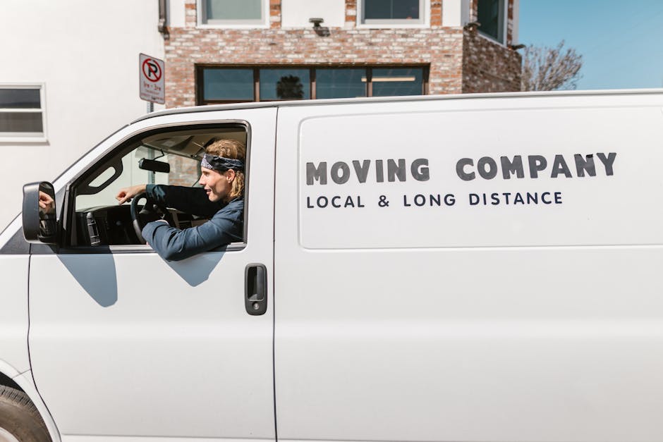A man with long blonde hair, wearing a blue jacket and a black headband, is sitting in the driver's seat of a white moving van, which has the words 'MOVING COMPANY' and 'LOCAL & LONG DISTANCE' printed on the side in large grey letters. The van is parked outside a residential property with a brick facade, multiple windows, and a clear sky in the background. The driver is holding the steering wheel with both hands, looking forward, preparing for a home relocation or furniture transport process. Inside, the van's interior is not fully visible, but it is implied to be equipped for packing and moving. Nearby pavement and a 'no parking' sign can be seen, indicating the vehicle's parking location during loading or unloading. The setting suggests the vehicle is positioned close to the property to facilitate efficient loading of boxes, furniture, and packaging materials wrapped in plastic or fabric blankets, typical in moving services provided by [COMPANY_NAME].