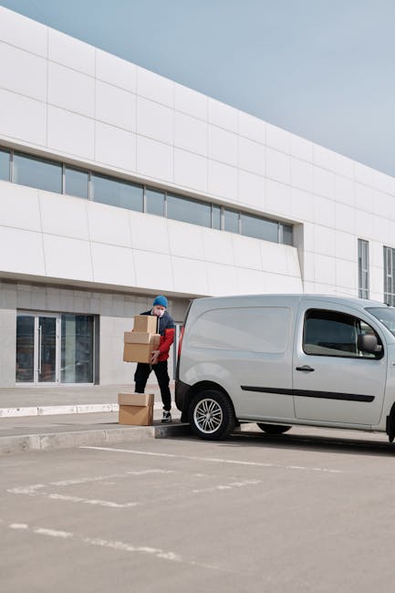 A man dressed in a red and black jacket, wearing a blue knit hat and a face mask, is standing outside a modern white building with large windows, lifting and stacking cardboard boxes near a small silver van parked on the paved pavement. The van is a compact commercial vehicle with black side mirrors, black wheel arches, and a sliding side door, positioned close to the building’s entrance. The scene captures a home relocation or furniture transport process, with the man preparing boxes for loading or unloading as part of a professional removals service provided by Man with Van Park Royal. The environment is well-lit with natural daylight, appearing to be a typical urban or business area facilitating packing and moving activities within the context of house removals or moving logistics.