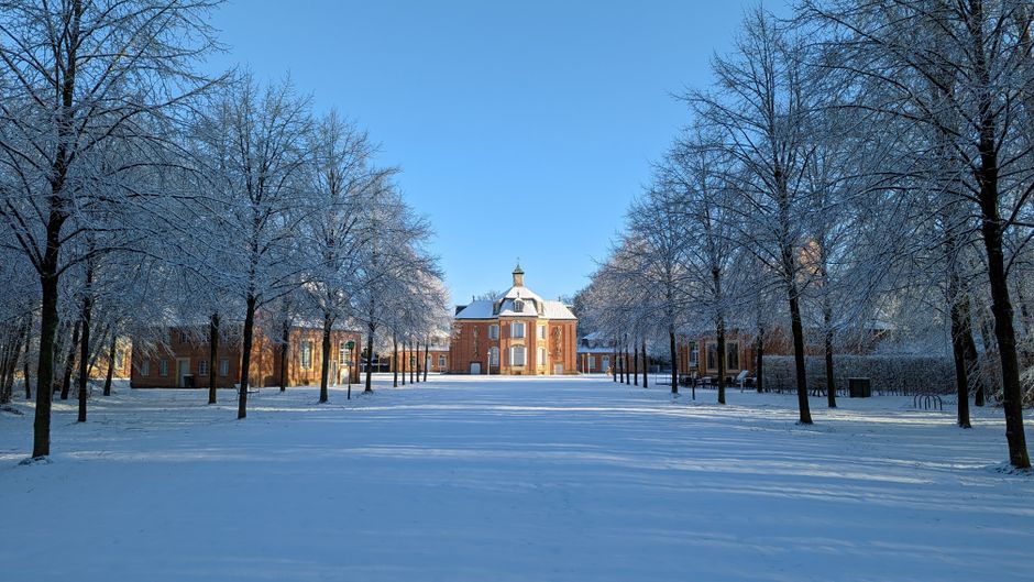 A modern two-storey house with a flat roof and a combination of brick, white, and wood cladding exterior, situated in a snow-covered landscape during winter. The house has large windows, some with bla